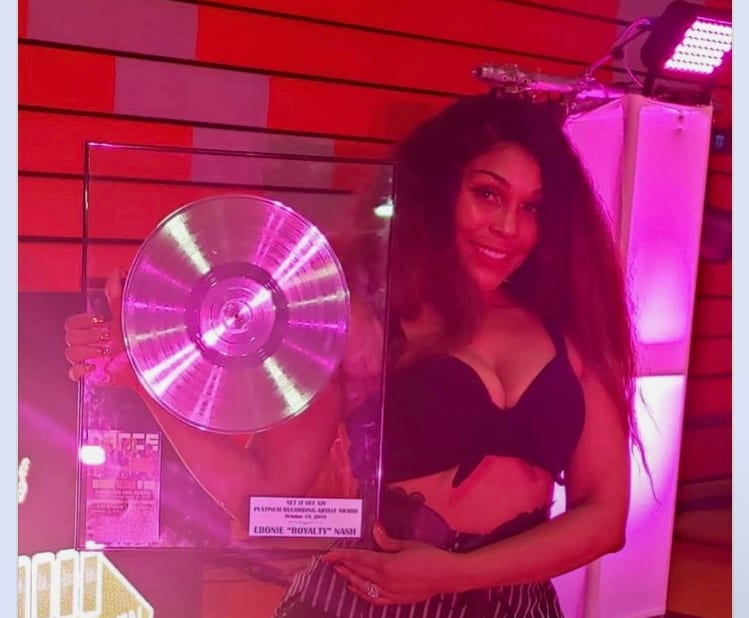 Standing beside a framed gold record award under pink light.