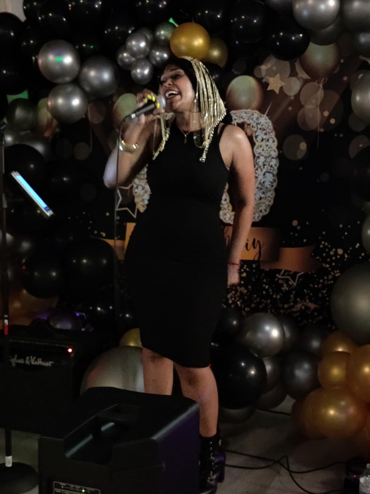 Performing in a black dress in front of a black and gold balloon arch.