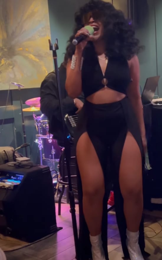Performing in black at a stand microphone, a large gold painting on the wall behind.
