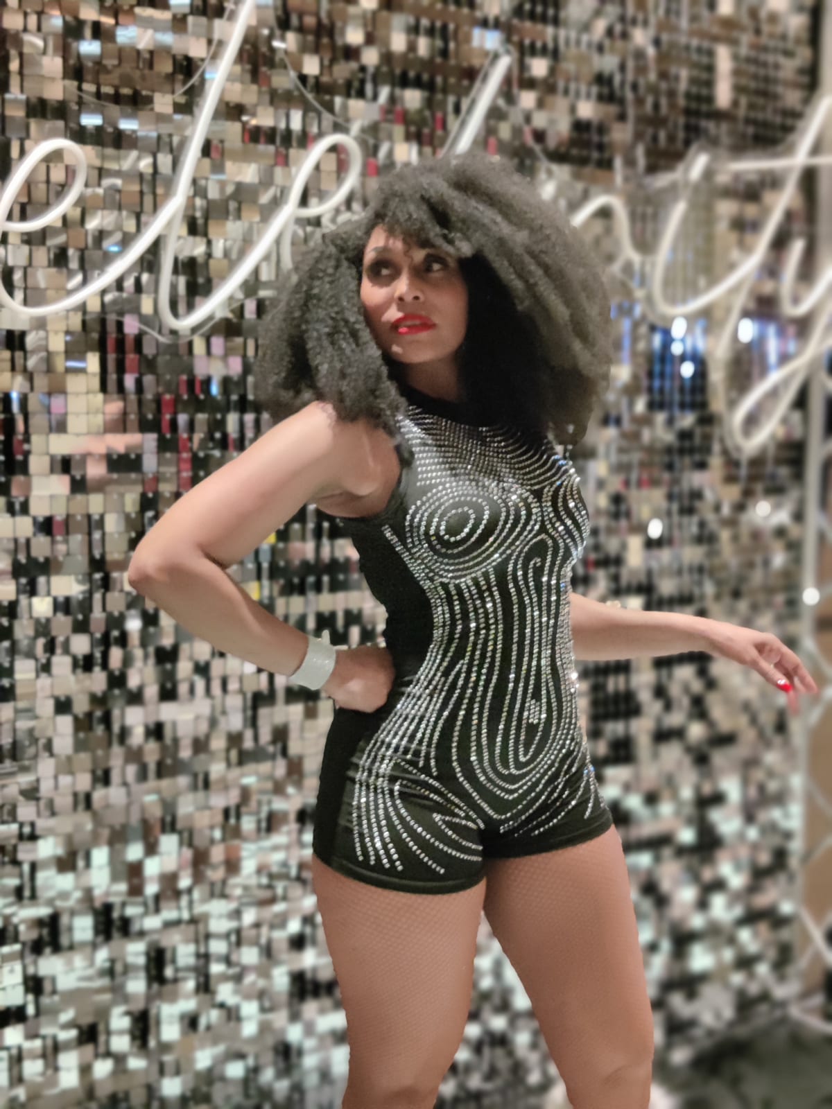 Posed in a sequinned bodysuit against a mirrored wall under a neon script sign.