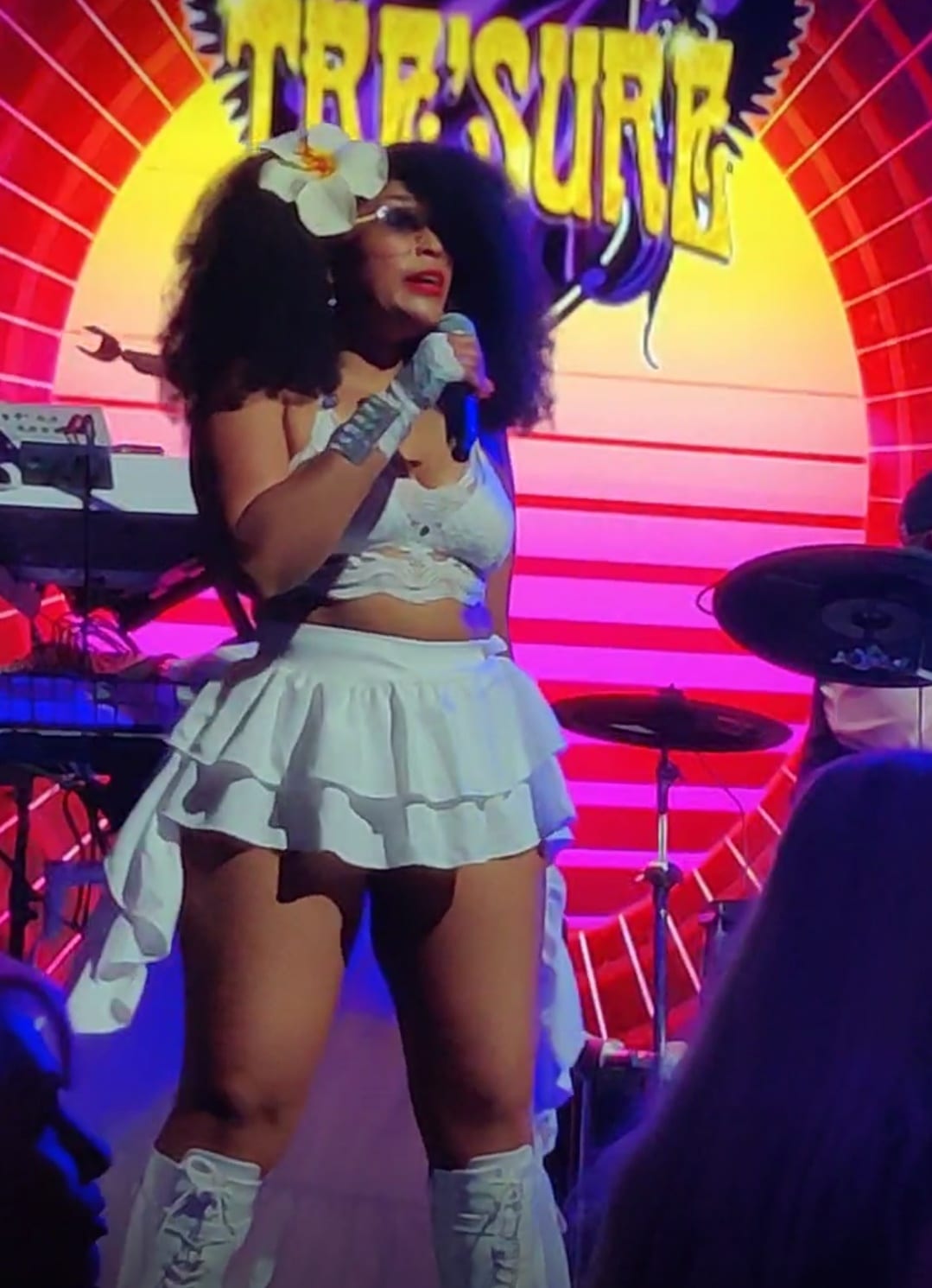 Singing in a white two-piece with a flower in her hair, in front of a red and pink neon arch.