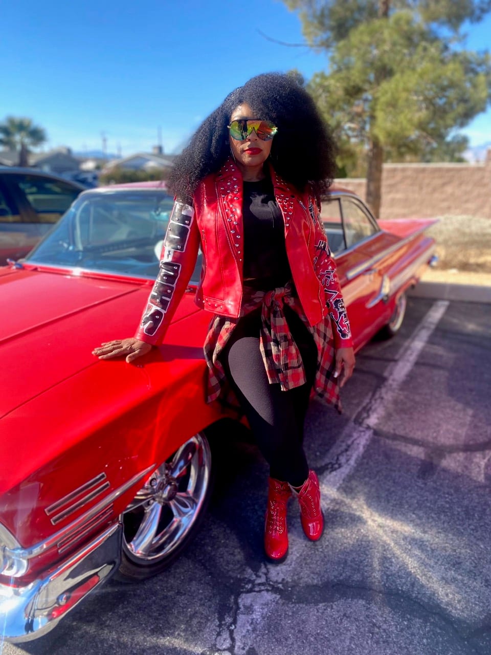 Leaning against a red classic car in a studded red jacket and sunglasses.