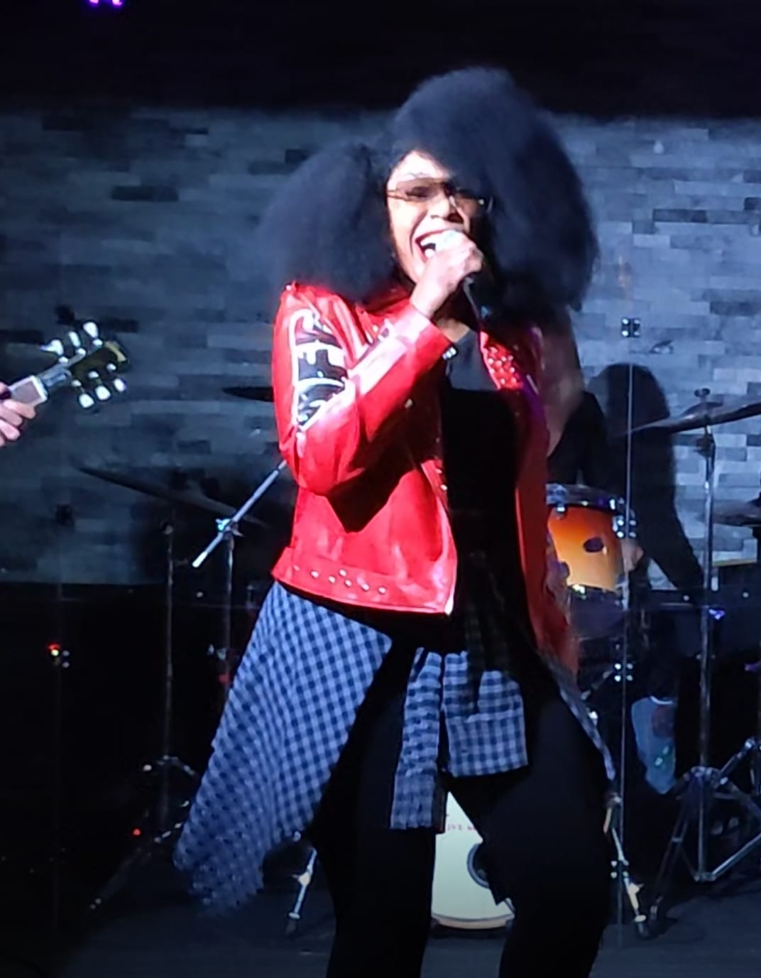 Laughing into the microphone in a red leather jacket against a grey stone wall.