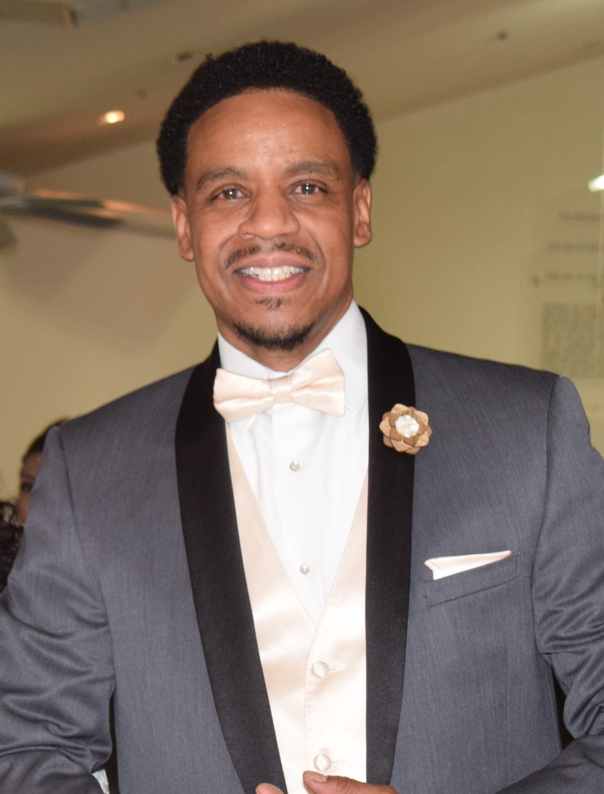 A man in a grey tuxedo with a white bow tie and a floral lapel pin.