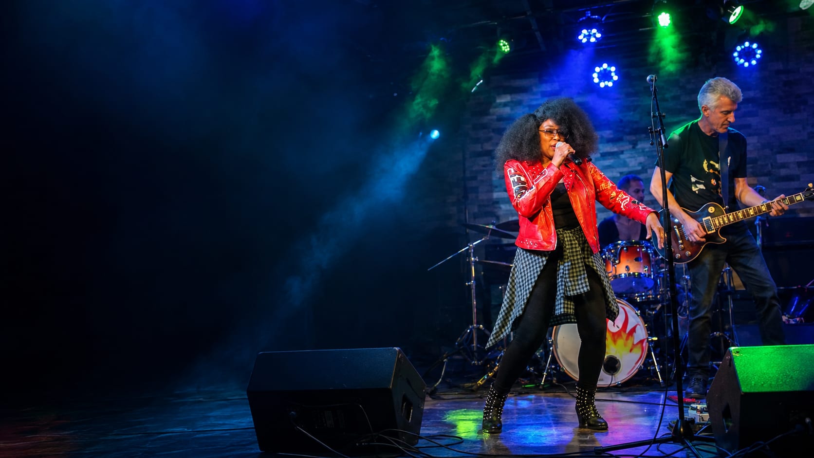 A singer in a red leather jacket performing on stage under blue and green lights, a guitarist and drum kit behind her.