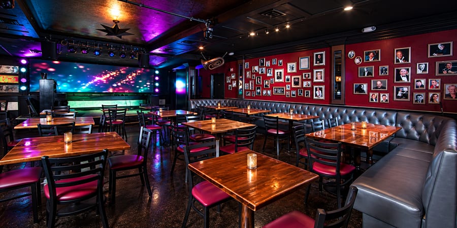 A supper club with cabaret tables and candles facing a lit stage, a wall of framed portraits alongside.